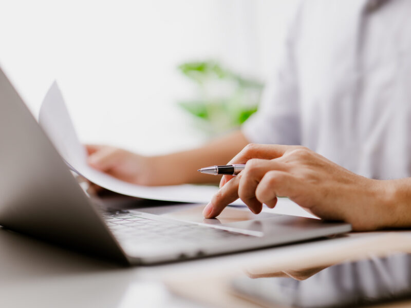 La main de l’homme d’affaires est sur le trackpad, tapant sur un clavier d’ordinateur portable, tout en lisant un document d’affaires.